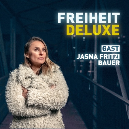 Jagoda Marinic in einem voluminösen, flauschigen Mantel trägt. Sie blickt leicht nach oben, mit einem nachdenklichen und zugleich hoffnungsvollen Ausdruck. Im Hintergrund erkennt man eine nächtliche, beleuchtete Fußgängerbrücke. „FREIHEIT DELUXE“ – „Freiheit“ in Weiß, „Deluxe“ in Gelb, was einen Kontrast erzeugt.